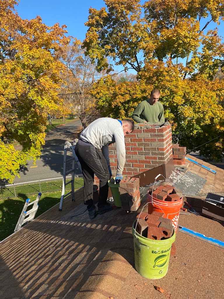 Two AAA Apex masons rebuilding a brick chimney on a shingle roof, with buckets of brick beside them