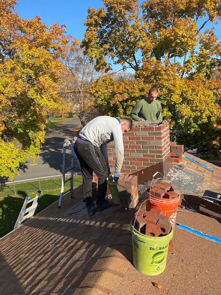 Two AAA Apex masons rebuilding a brick chimney on a shingle roof, with buckets of brick beside them